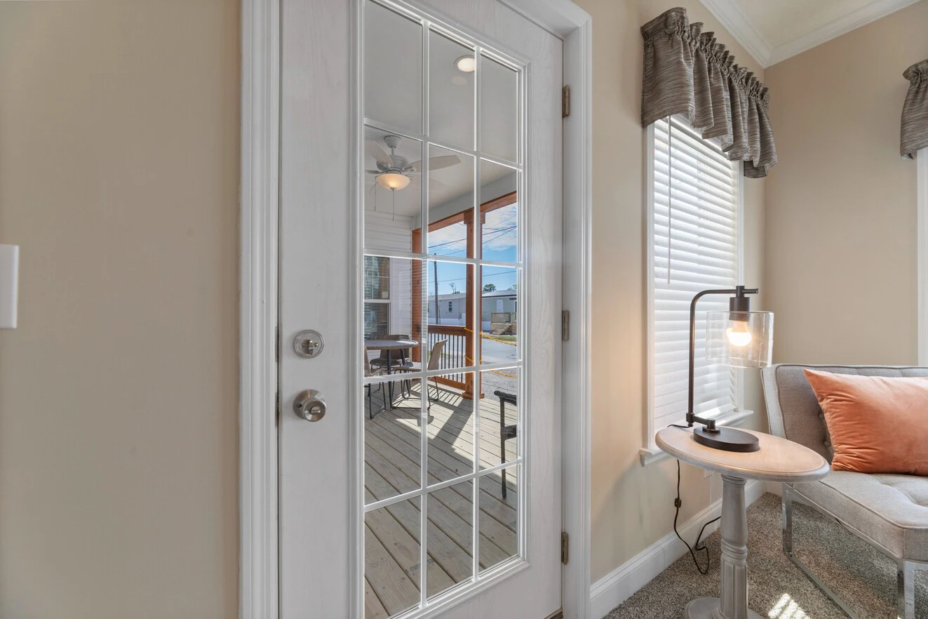 Glass-paneled door with white frame leads to a covered patio; inside, a gray couch with an orange pillow and a round table with a modern lamp are visible.