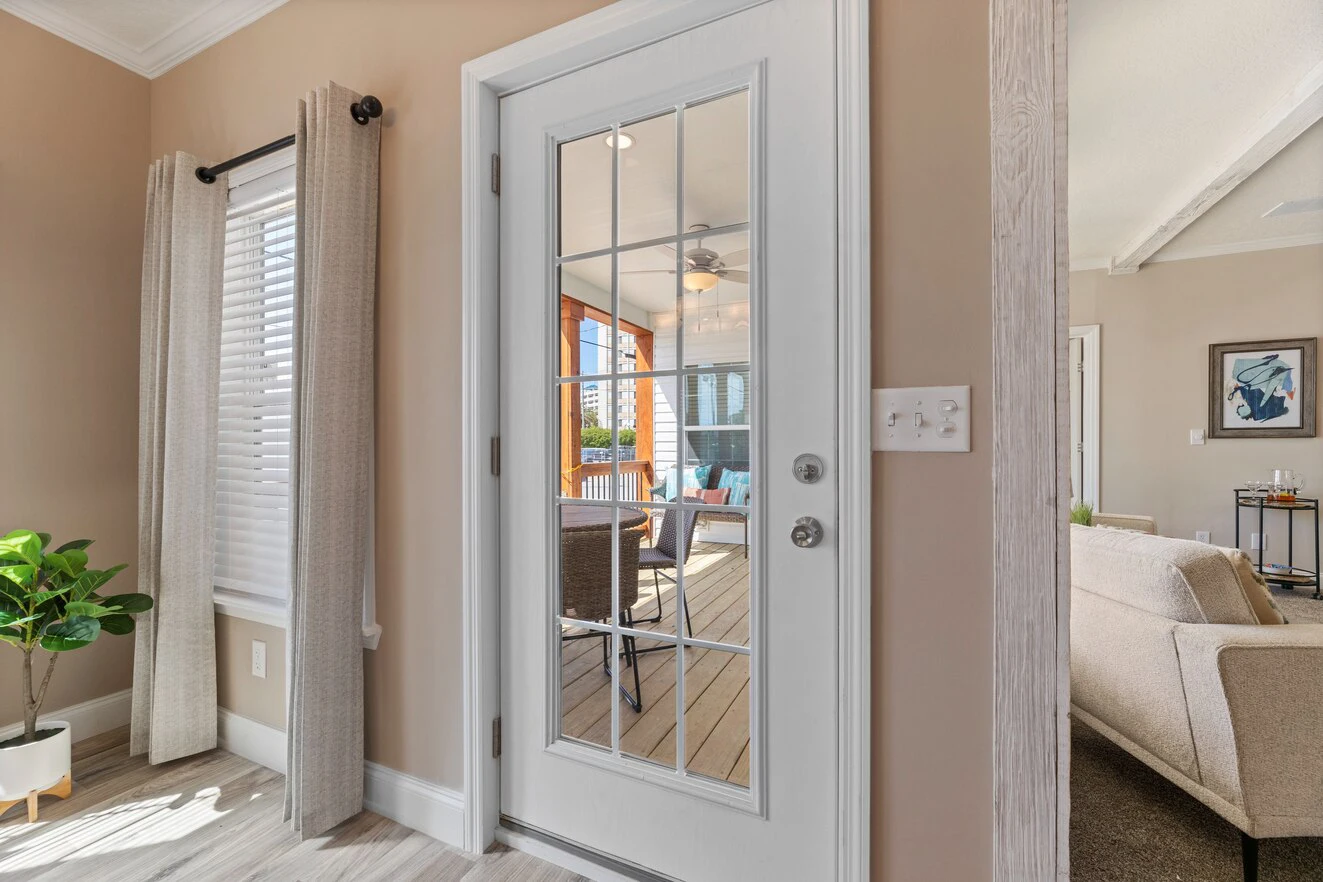 A glass-paneled door with a silver handle leads to a sunroom with wicker chairs; beige walls and a window with curtains are in the foreground.