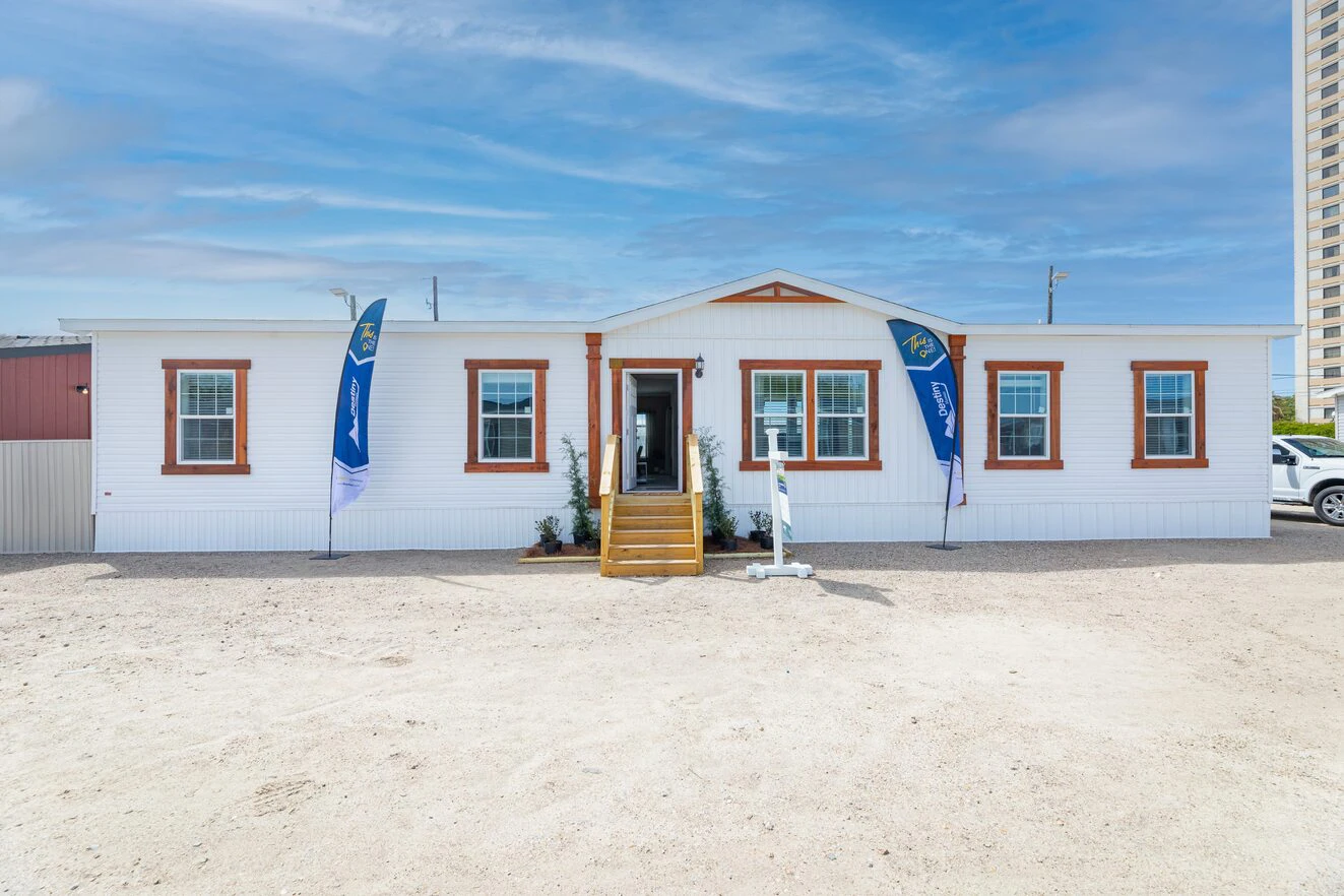 A single-story prefabricated home with white siding, brown trim, front steps, and blue flags on either side, set on a gravel lot under a partly cloudy sky.