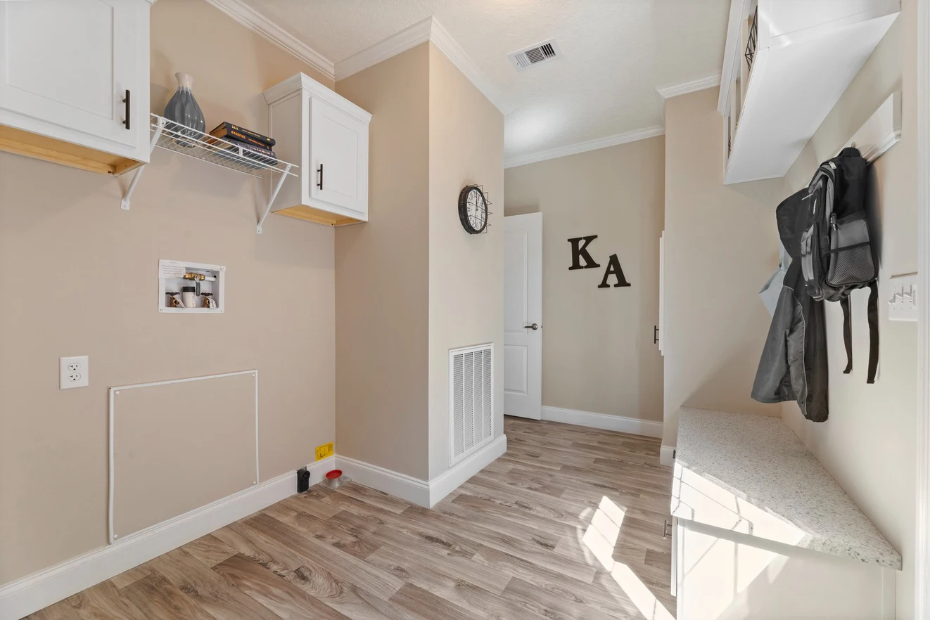 Laundry room with light wood floors, upper cabinets, a wall clock, hooks with backpacks, and a countertop. Shelving above the washer/dryer space holds decor and cleaning items.
