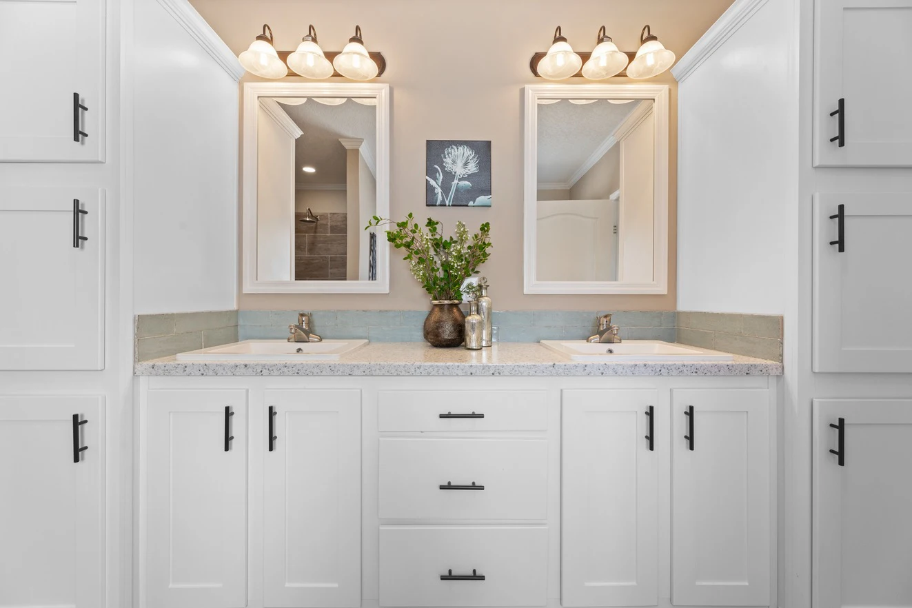 Double bathroom vanity with white cabinets, two mirrors, light fixtures above each, and a vase with greenery in the center. A small framed picture hangs between the mirrors.