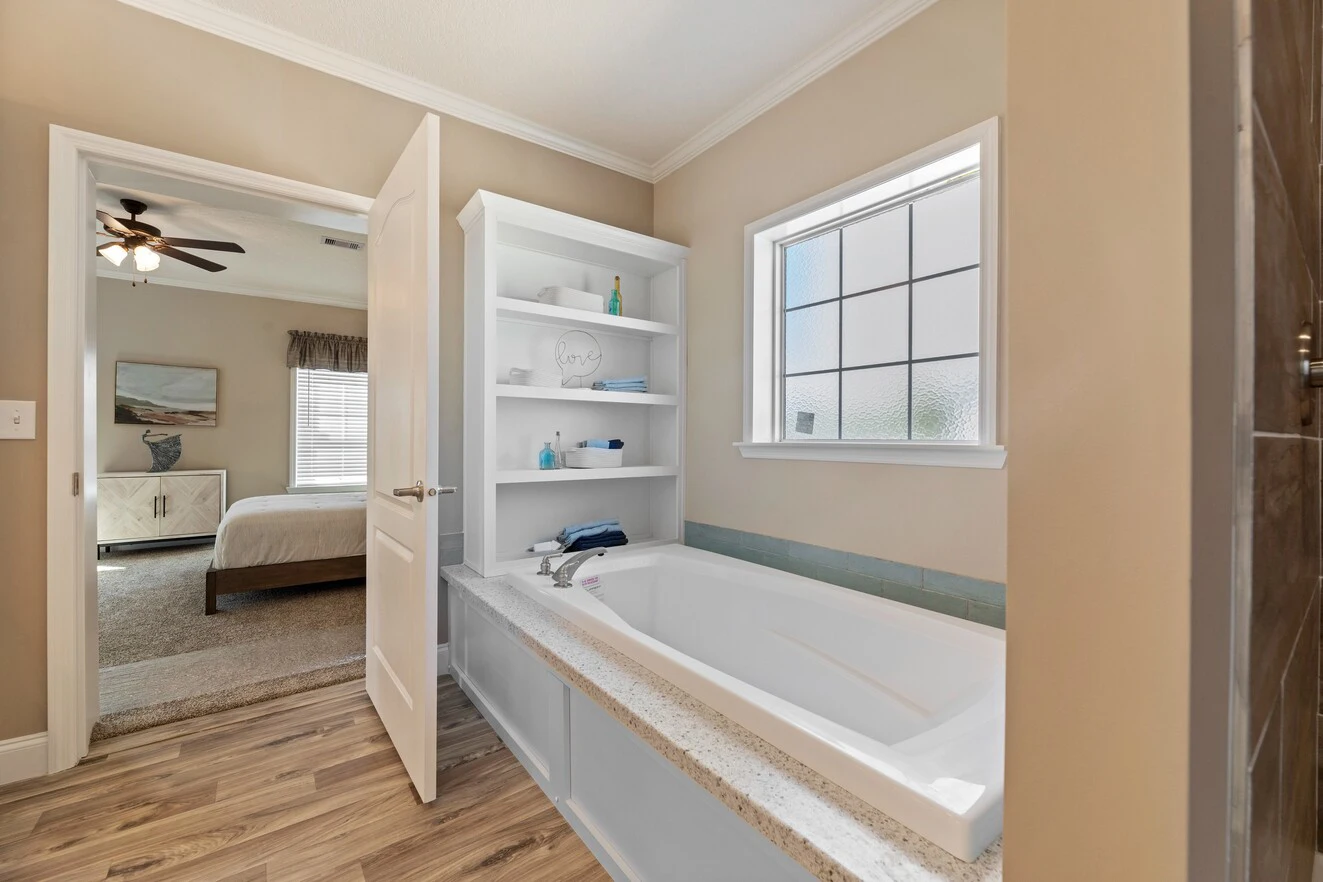 Bathroom with a built-in bathtub, open shelving, frosted window, and view into a bedroom with a ceiling fan and bed.