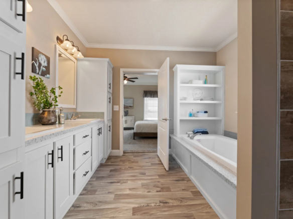 Modern bathroom with white cabinets, a built-in bathtub, plant decor, wood-look flooring, and a view into a bedroom through an open door.
