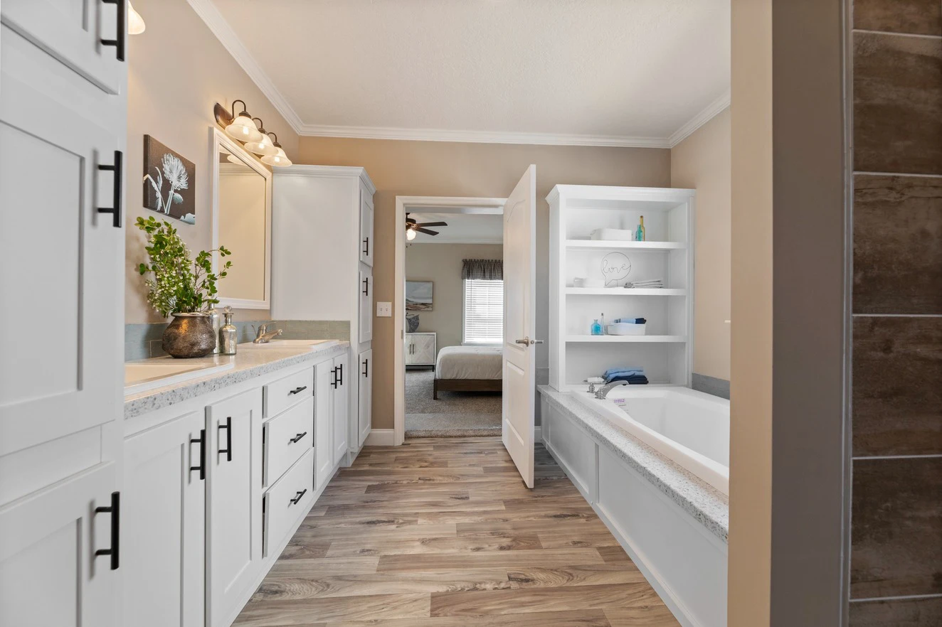Modern bathroom with white cabinets, a built-in bathtub, plant decor, wood-look flooring, and a view into a bedroom through an open door.