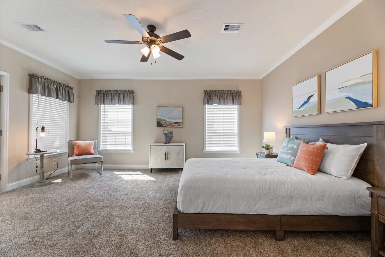 Spacious bedroom with beige walls, carpeted floor, large bed, two windows with gray valances, ceiling fan, and minimalist decor.