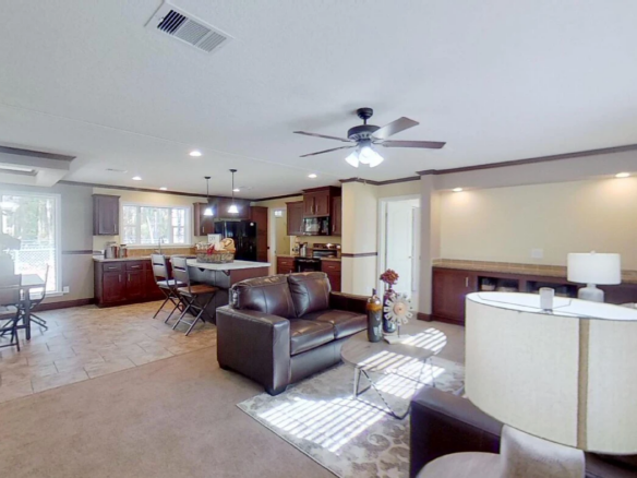 Open-concept living area with a kitchen, dining table, and sitting area featuring brown sofas, ceiling fans, and large windows letting in natural light.