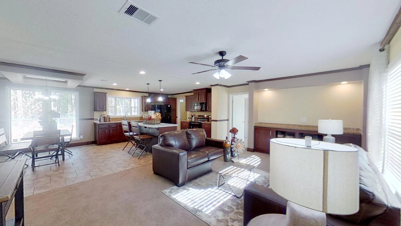 Open-concept living area with a kitchen, dining table, and sitting area featuring brown sofas, ceiling fans, and large windows letting in natural light.