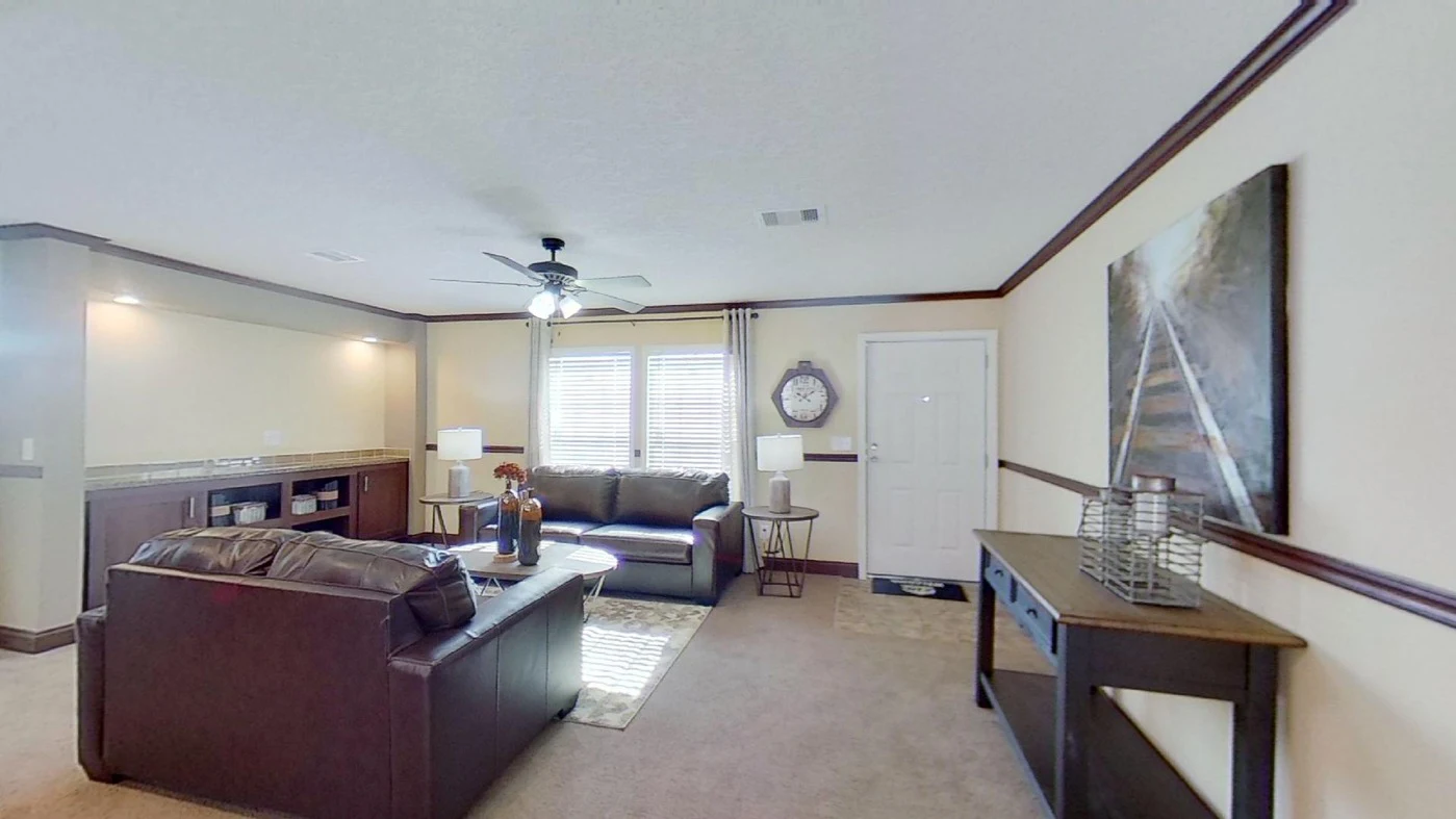 A living room with brown leather sofas, a ceiling fan, a large window with blinds, a wall clock, and a console table with decor items.