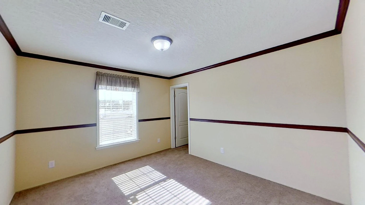 Empty room with beige walls, brown trim, carpeted floor, a window with blinds and valance, ceiling light fixture, and a closed white door. Sunlight casts shadows on the floor.