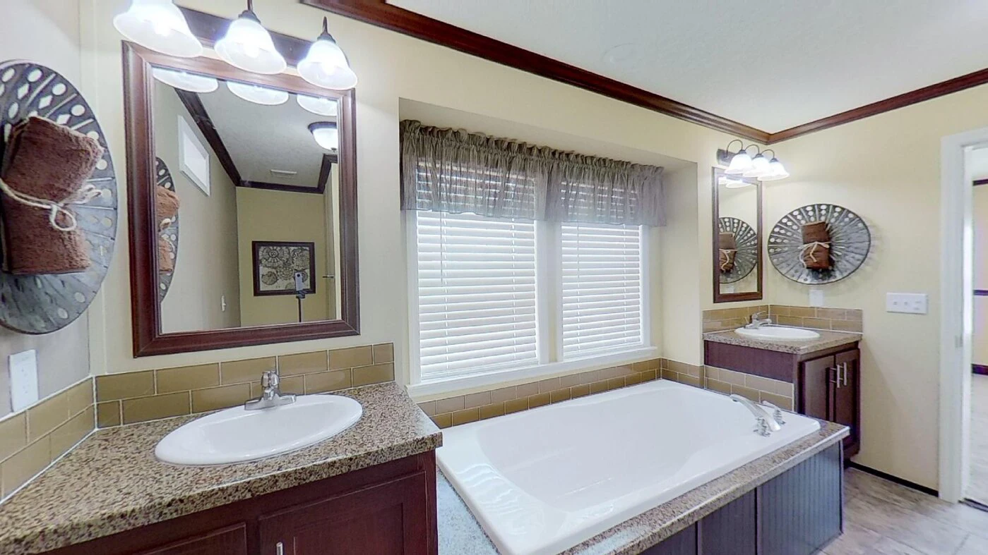 Bathroom with a large white bathtub between two sinks, wall mirrors above each sink, and large windows with sheer curtains letting in natural light.