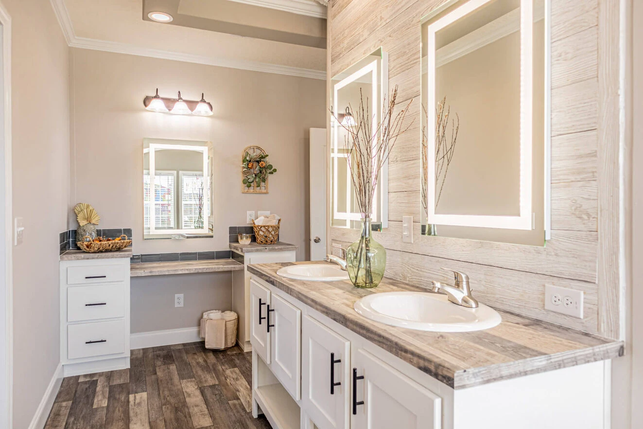 Modern bathroom with double sinks, illuminated mirrors, decorative branches in a glass vase, wood-look flooring, and a vanity area with a cushioned stool.
