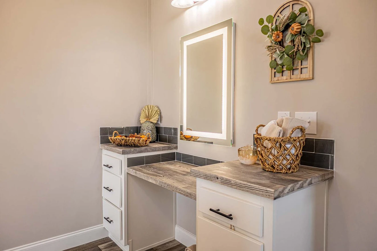 A vanity with white cabinets, a wooden countertop, an illuminated mirror, and decorative baskets stands against a light-colored wall with a floral wall hanging.