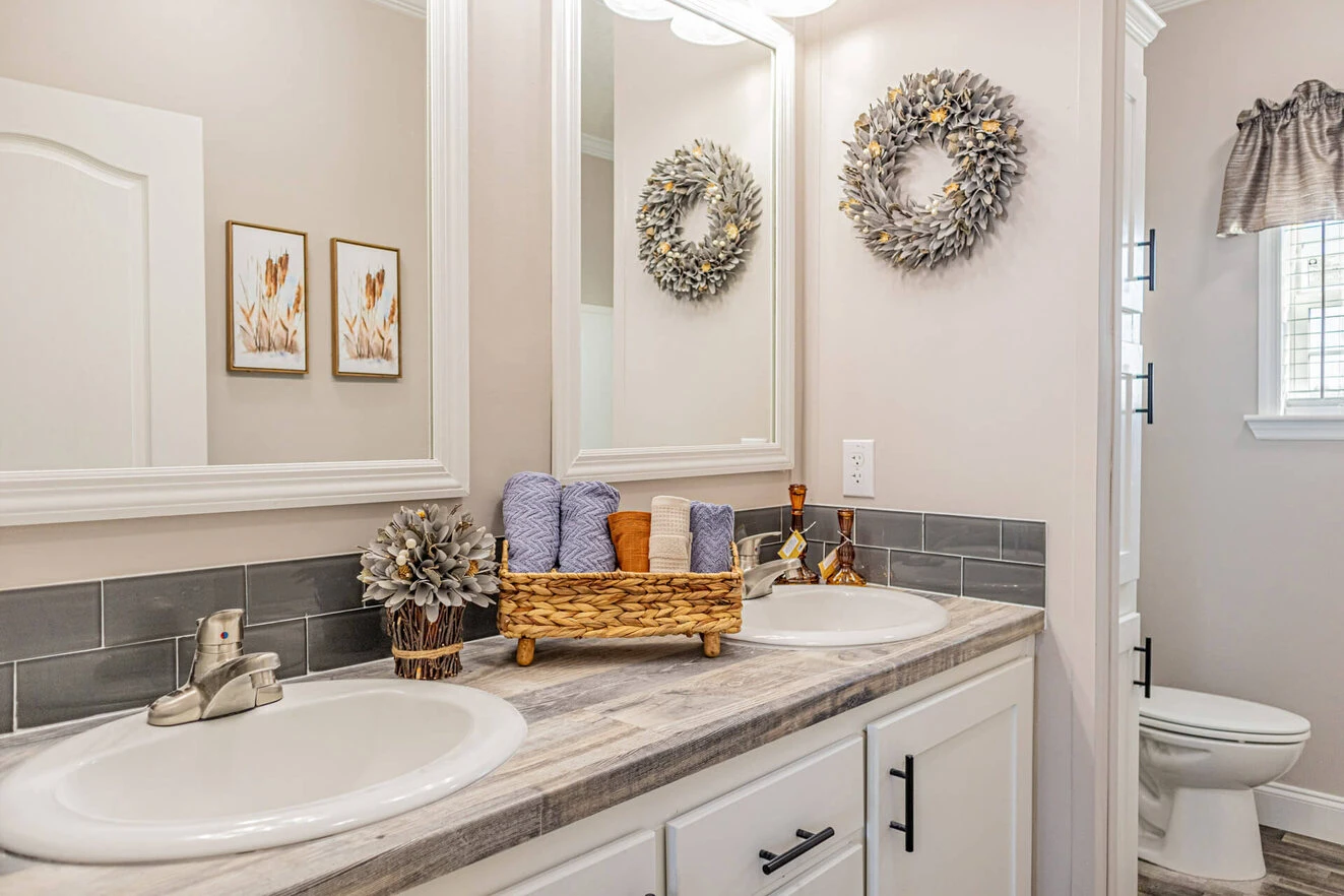 Double bathroom vanity with gray backsplash, two sinks, a basket of towels, decorative wreaths on the wall, and a toilet visible in the background.