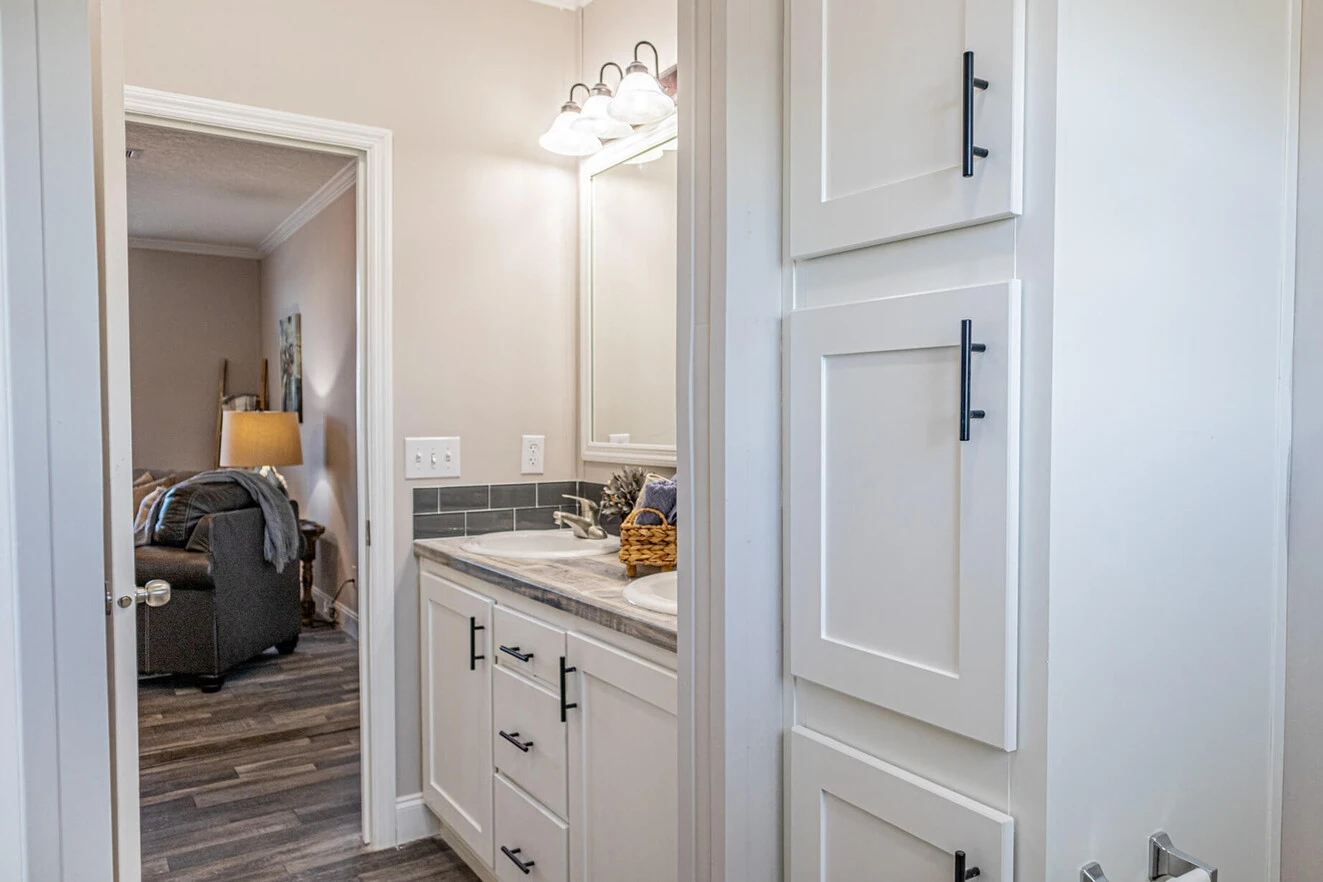 Bathroom with white cabinets, marble countertop, and a mirror; view through the open door shows a living room with an armchair and floor lamp.