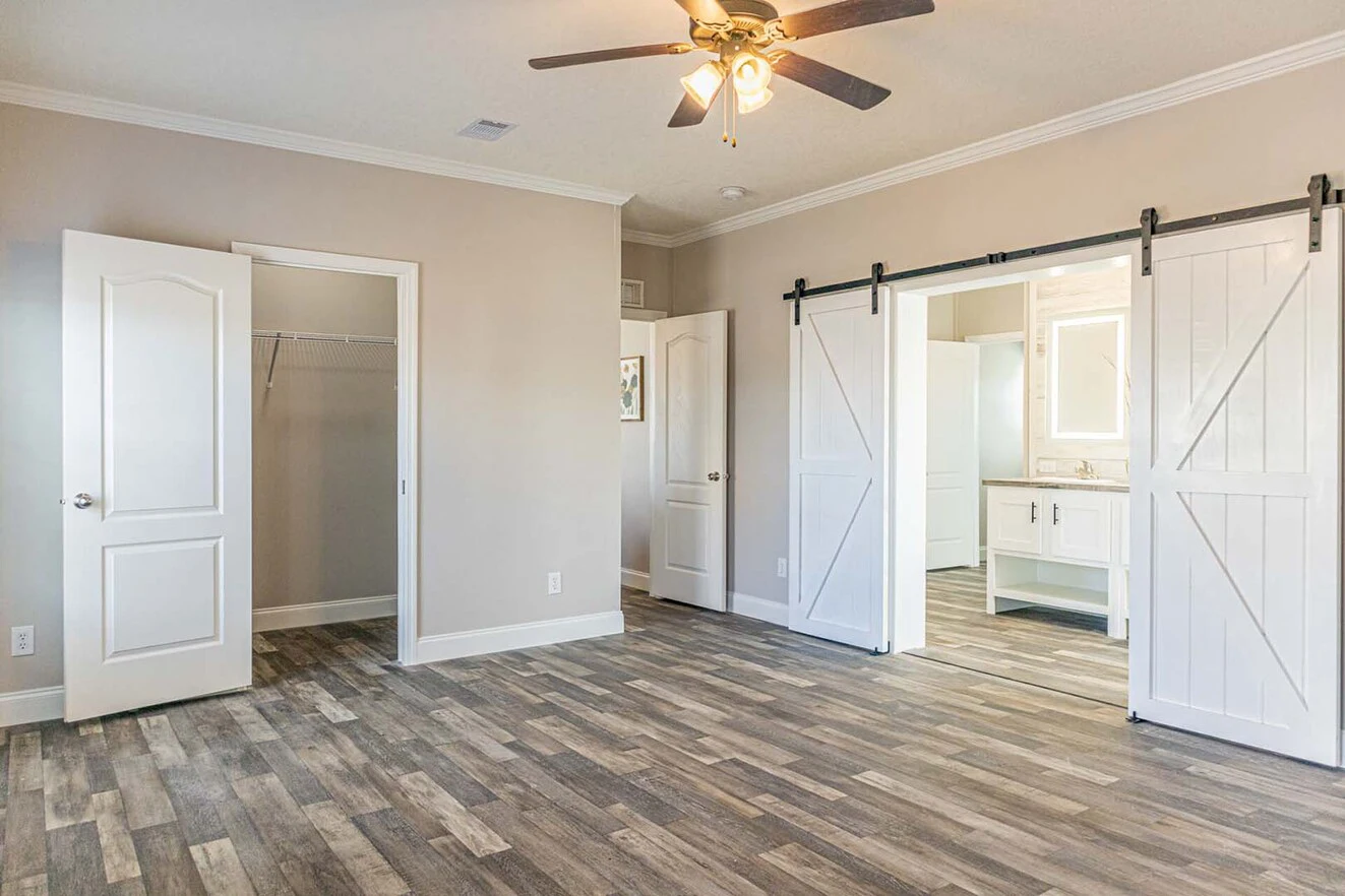 Spacious empty bedroom with wood-look flooring, a ceiling fan, open closet door, and sliding barn doors leading to a bathroom.