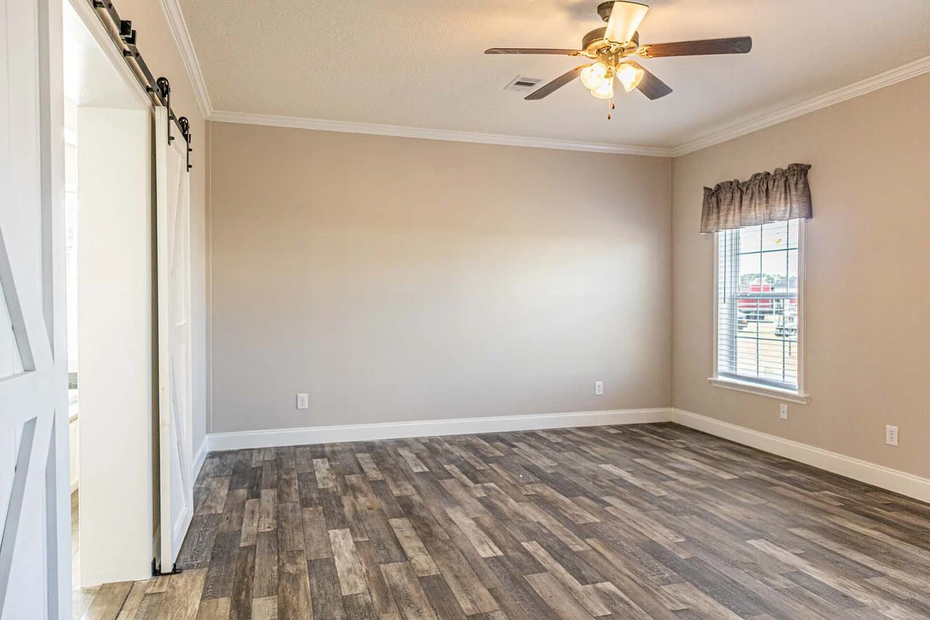 Empty room with wood-look flooring, beige walls, a ceiling fan with lights, a window with valance, and a sliding barn door partially open to another room.