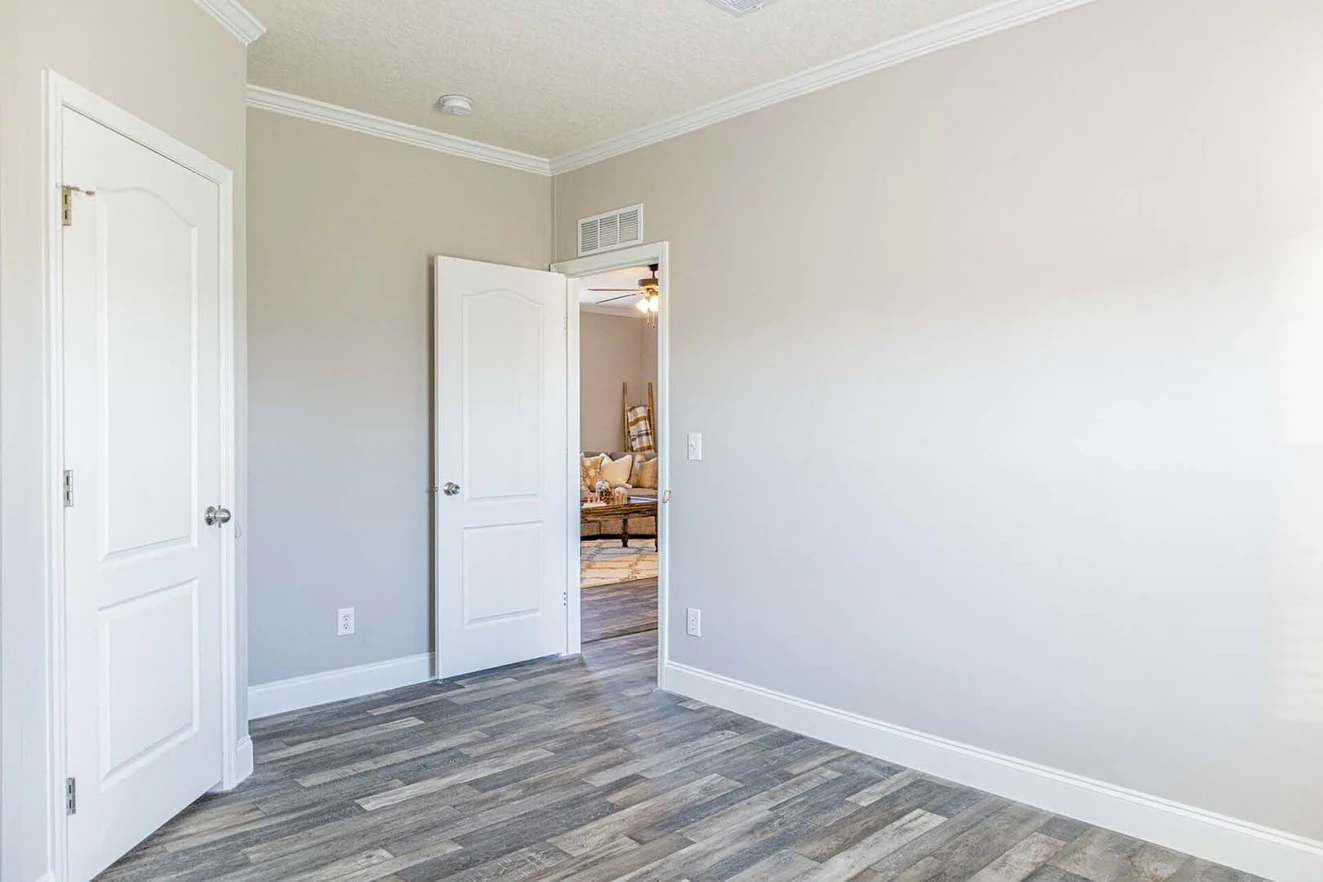 Empty room with light gray walls, white trim, and wood-style flooring. An open door reveals a glimpse of a furnished living area.