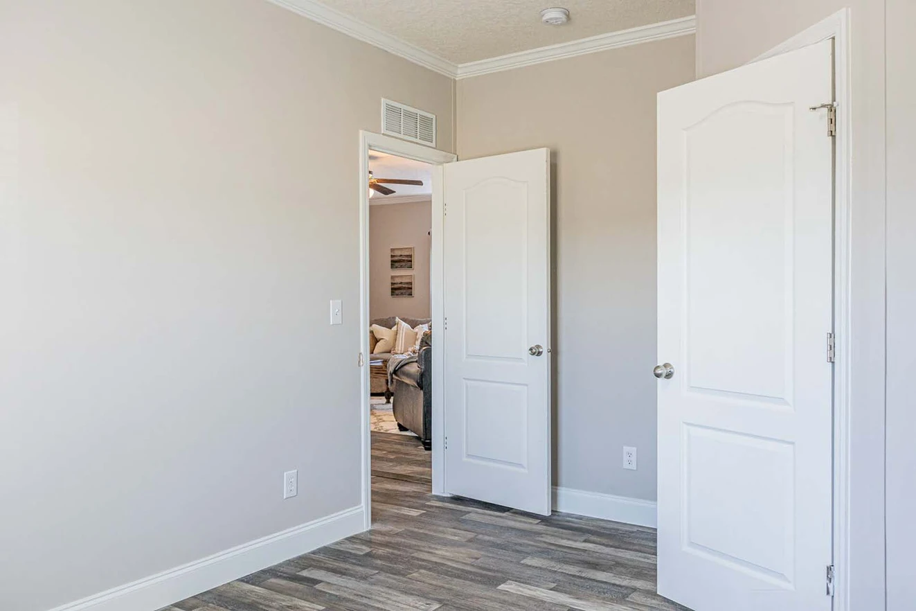 Empty room with light-colored walls and wood-patterned flooring, two white doors open, one leading to a furnished living space.