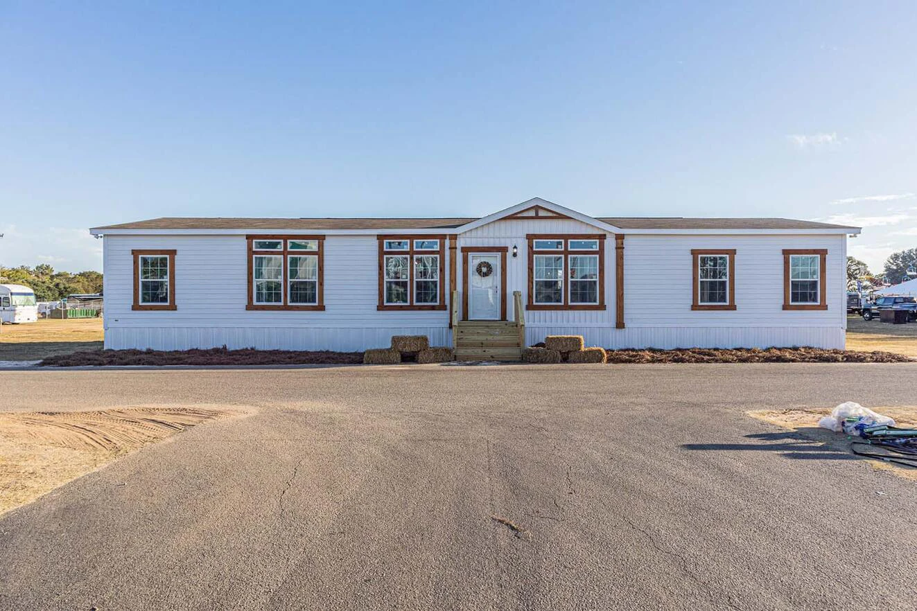 A single-story white manufactured home with brown trim, a central front door, and steps leading up from a paved road, with hay bales and open land nearby.