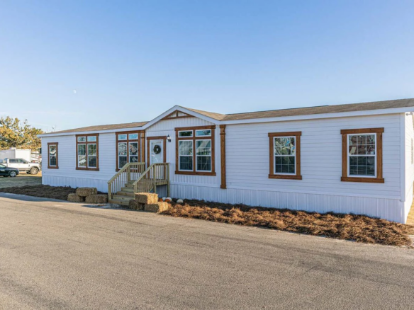 A white manufactured home with brown trim sits next to a paved road, featuring several windows and a small front porch with steps and a wreath on the door.