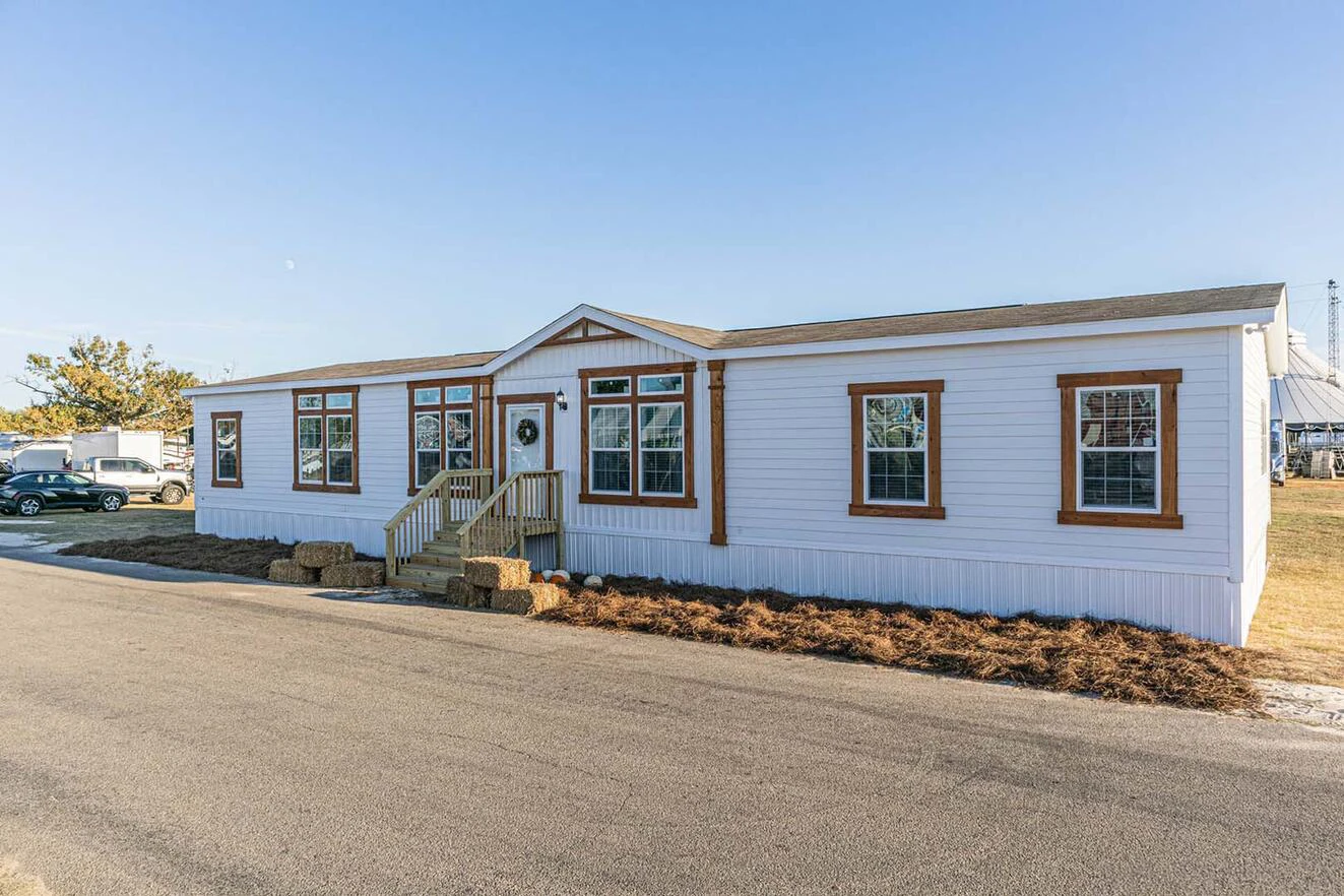 A white manufactured home with brown trim sits next to a paved road, featuring several windows and a small front porch with steps and a wreath on the door.
