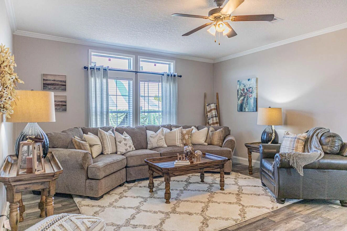 A cozy living room with a sectional sofa, armchair, two lamps, a coffee table, and large windows with light streaming in. Neutral tones and decorative accents complete the space.