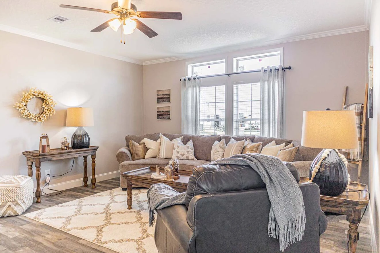 Bright living room with neutral decor, a sectional sofa, armchair, wooden coffee table, and side tables with lamps. Large windows with sheer curtains let in natural light.