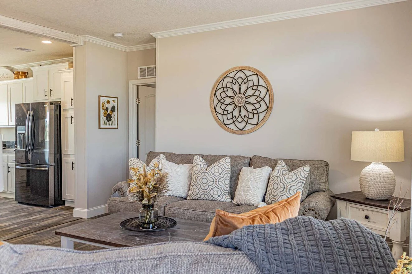 A living room with a gray sofa, assorted pillows, a round wall decor, a side table with a lamp, and a coffee table with a floral arrangement; kitchen visible in the background.