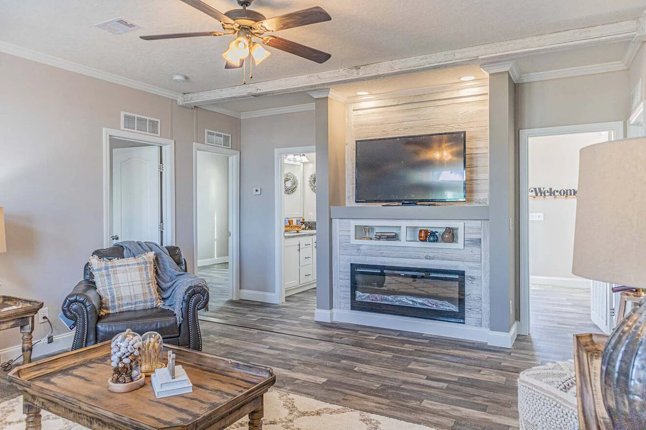 A living room with a ceiling fan, wall-mounted TV above a fireplace, armchair, wooden coffee table, and wood flooring. Doors lead to adjacent rooms.