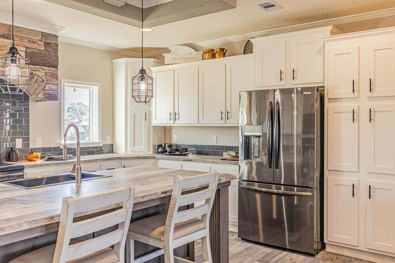 Modern kitchen with white cabinets, stainless steel refrigerator, marble island with sink, two wooden chairs, and hanging pendant lights.