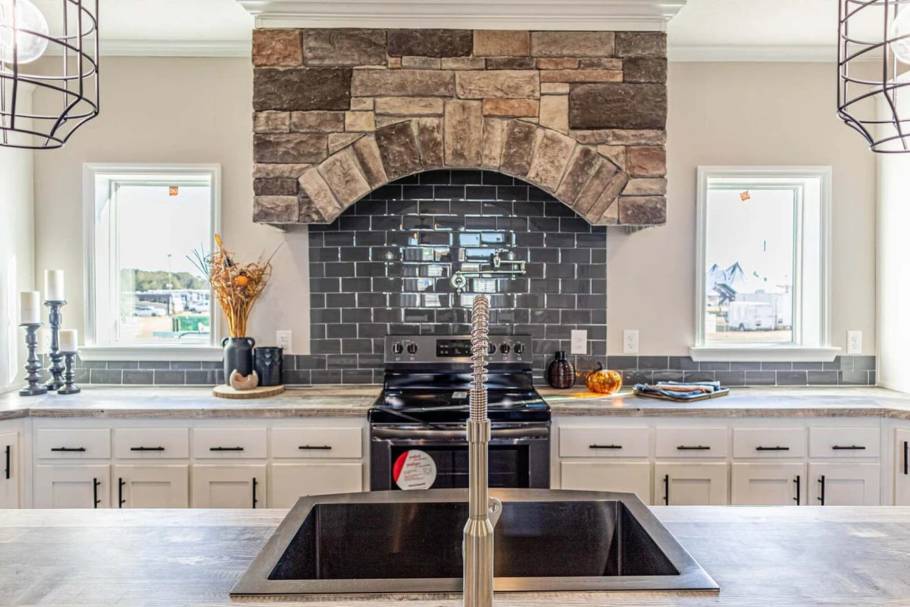 Modern kitchen with a central black sink, stainless steel faucet, black stove, gray subway tile backsplash, stone range hood, and two windows on either side.