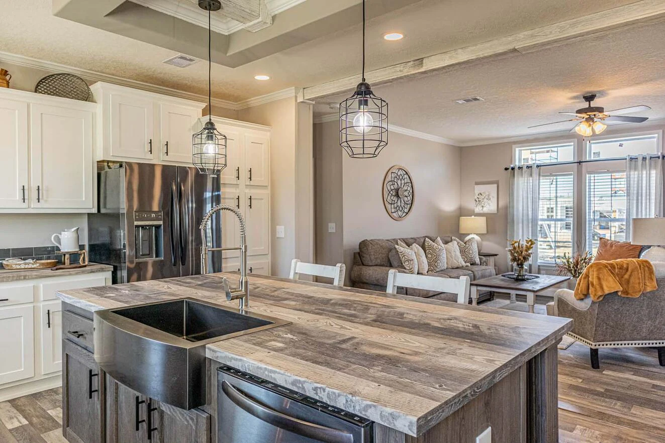 Modern kitchen with a farmhouse sink and wood island overlooks a living room with gray sofa, armchair, round clock, and large windows with curtains.