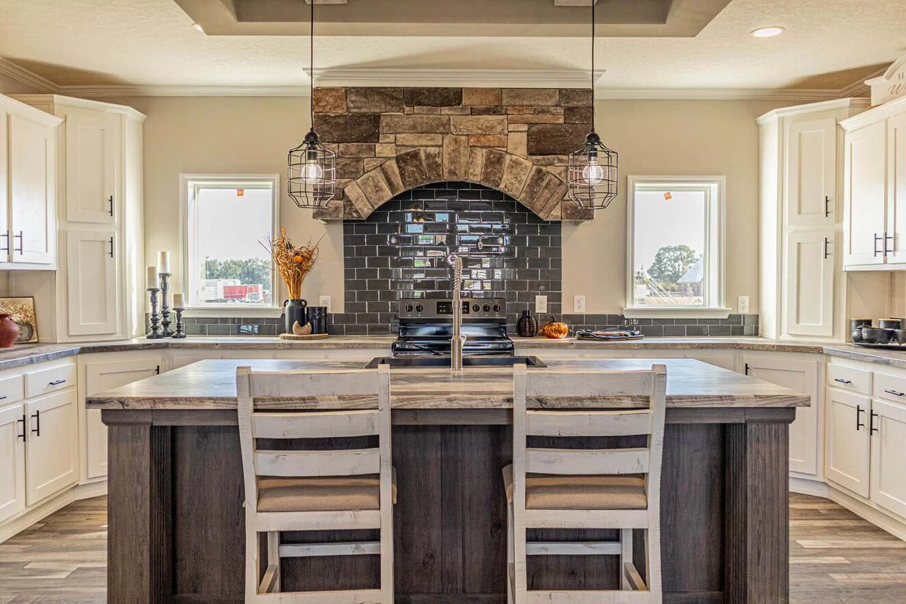 Modern kitchen with a large island, two chairs, white cabinetry, pendant lights, and a stone and tile accent above the stove. Natural light comes through two small windows.