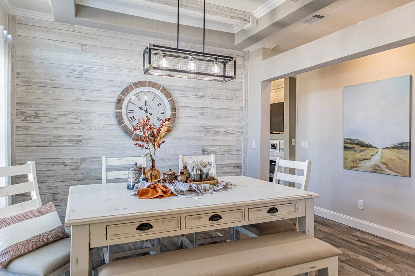 A bright dining room with a white rustic table, matching chairs, a bench, wall clock, fall-themed decor, and a modern black light fixture. A landscape painting hangs on the wall.