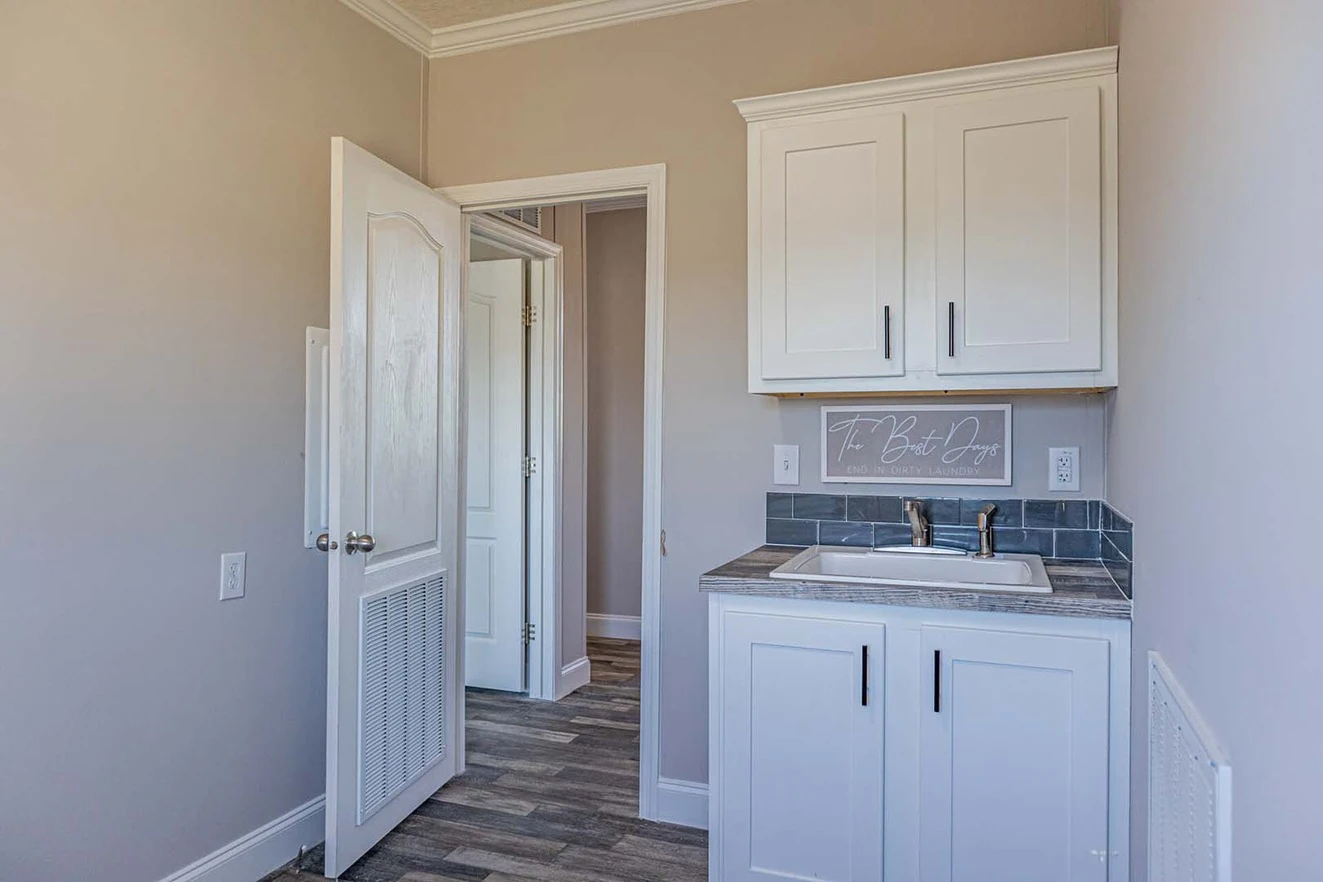 A small utility room with white cabinets, a sink, gray tile backsplash, and an open door leading to a hallway with wood-patterned flooring.