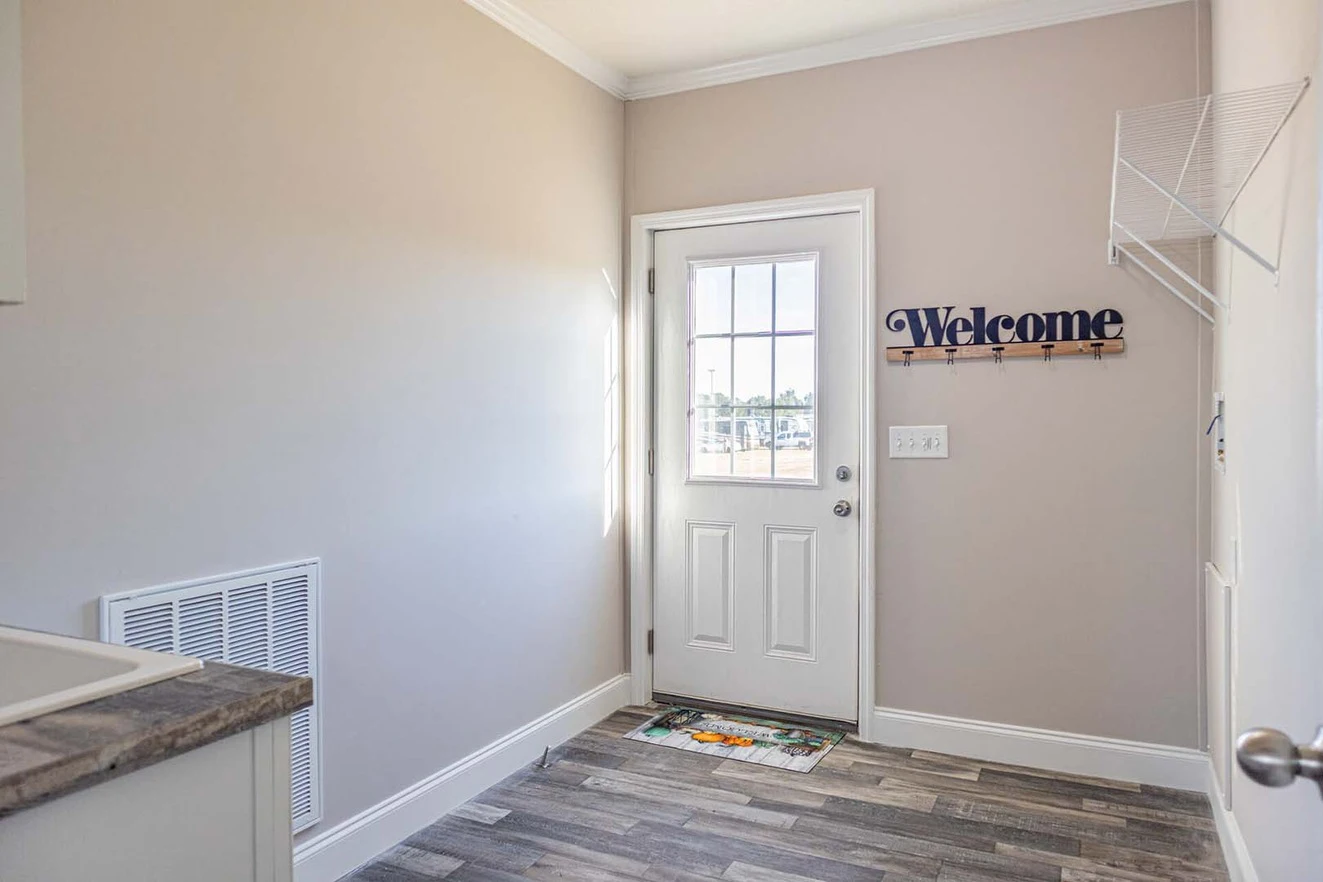 A small entryway with a door featuring a window, a welcome sign with hooks on the wall, a vent, a doormat, and light-colored walls and flooring.