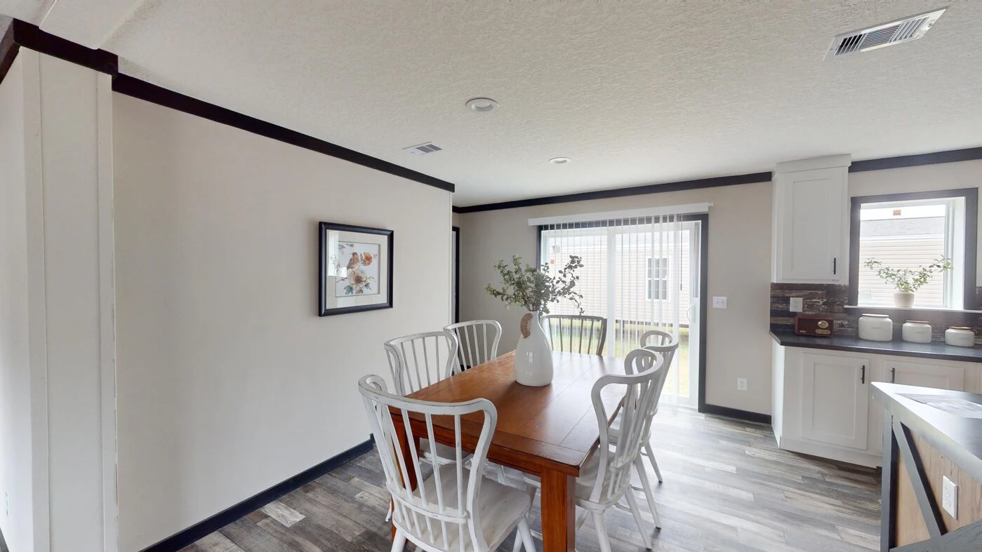 A dining area with a wooden table, six white chairs, a white vase with branches, wall art, large window, and sliding glass door leading outside.