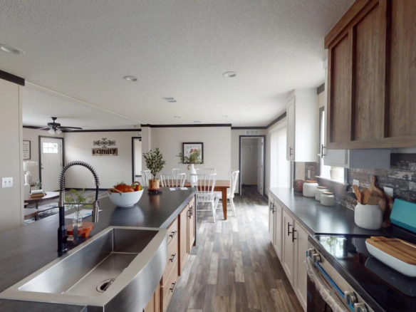 Modern kitchen with an island sink, fruit bowl, and dining area in the background. Wood accents and stainless steel appliances are visible throughout the space.