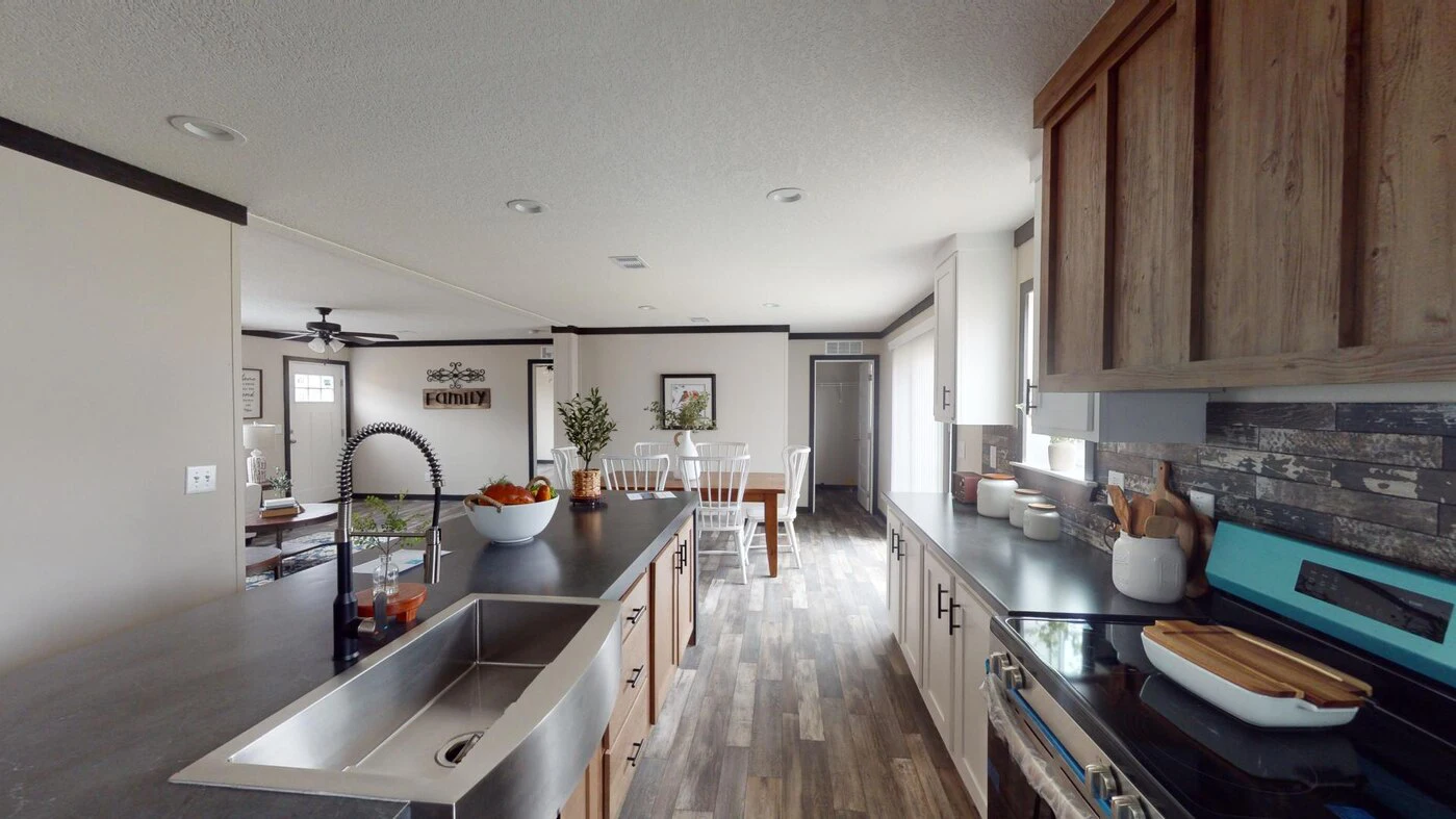 Modern kitchen with an island sink, fruit bowl, and dining area in the background. Wood accents and stainless steel appliances are visible throughout the space.