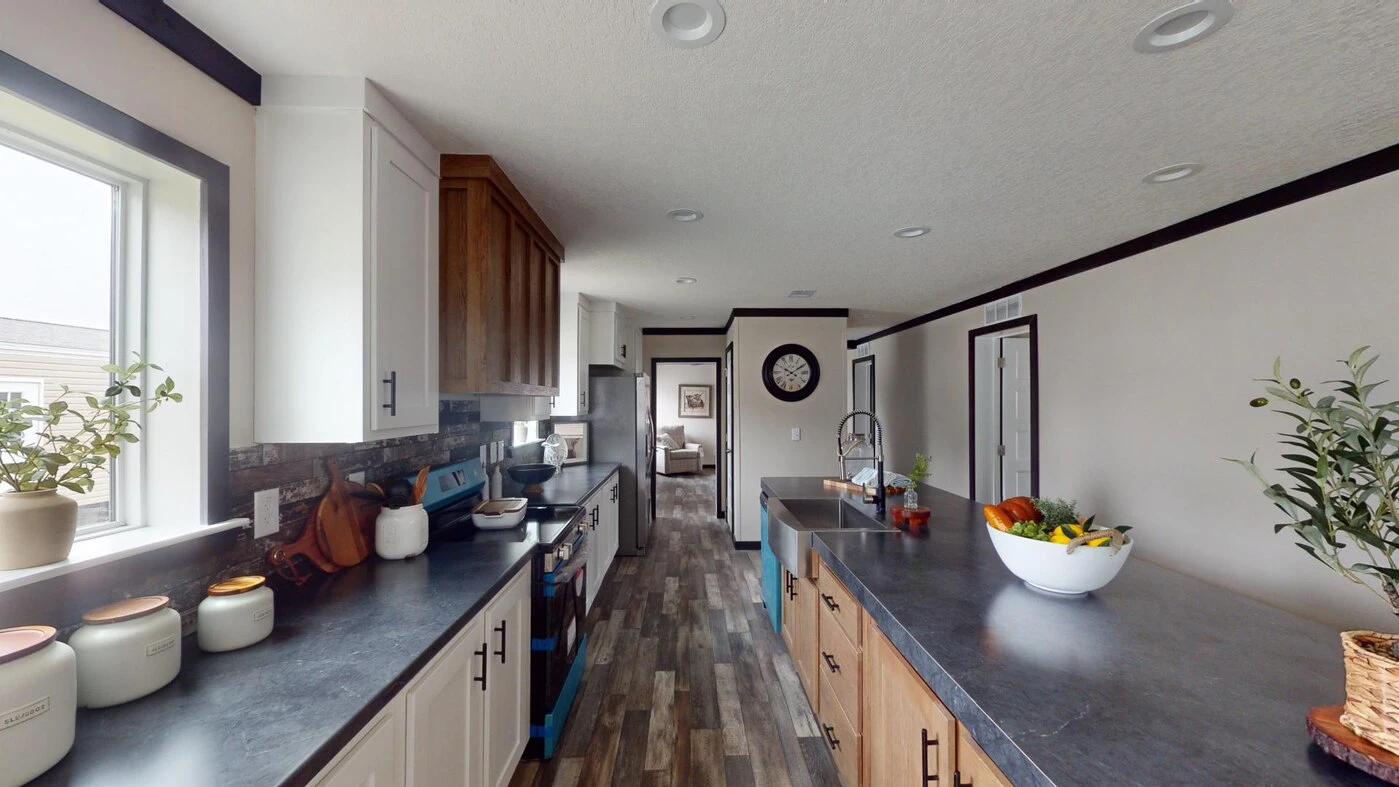 Modern kitchen with dark countertops, wooden cabinets, potted plants, a bowl of fruit, and a clock on the far wall. Natural light enters through a window on the left.