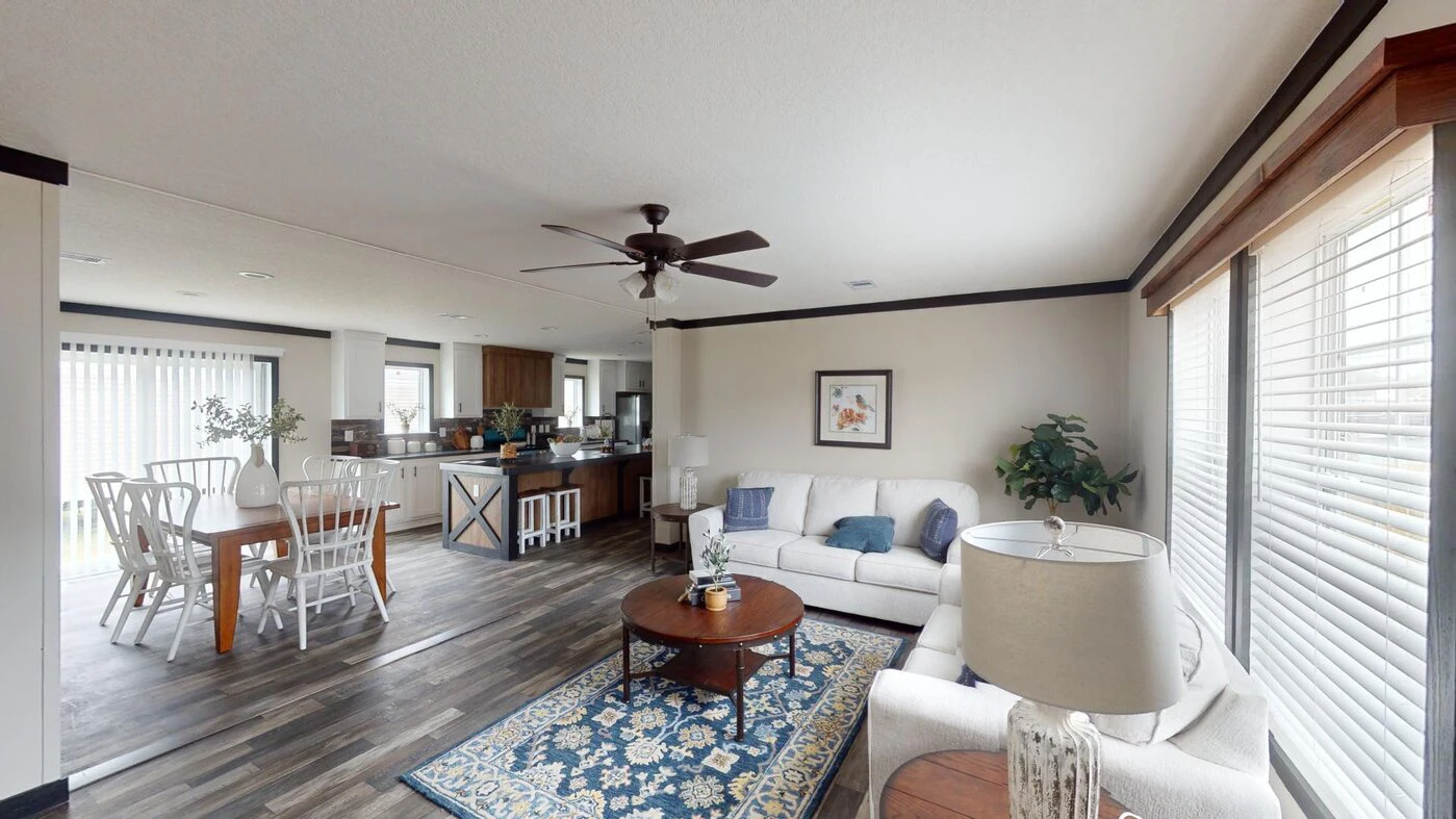 Open-plan living and dining area with white furniture, round wooden coffee table, rug, large windows with blinds, ceiling fan, and kitchen in the background.