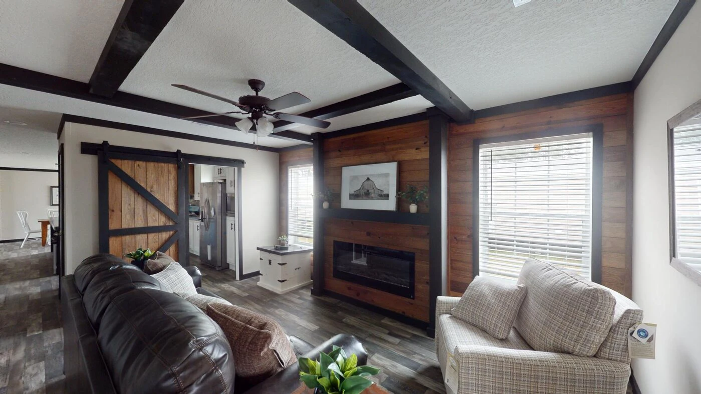 A living room with a dark leather sofa, plaid armchair, ceiling fan, barn door, fireplace with wood accents, and large windows with blinds.