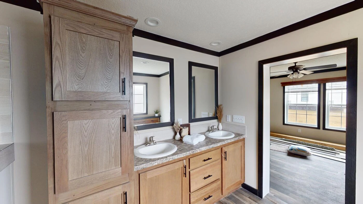 A bathroom with a double sink vanity, two mirrors, light wood cabinets, and a view into an adjacent room with a ceiling fan and large windows.