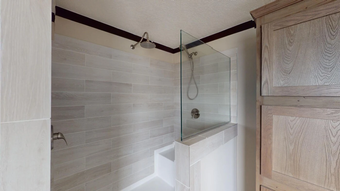 Walk-in shower with beige tile walls, partial glass divider, built-in bench, and a mounted showerhead. A wooden cabinet is visible to the right.