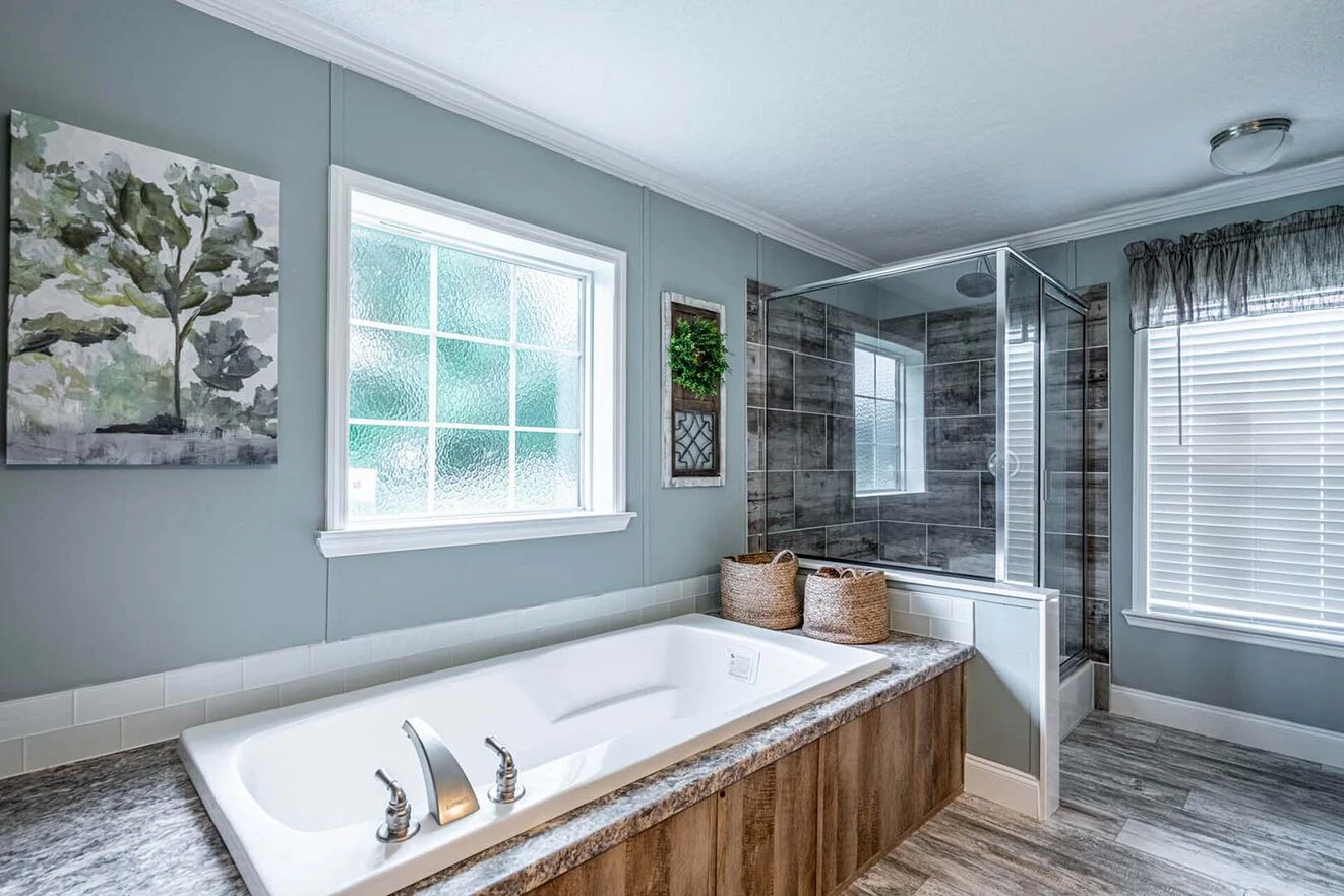 Modern bathroom with a white bathtub, wood-accented base, glass-enclosed shower, two windows, and decorative wall art with a plant.