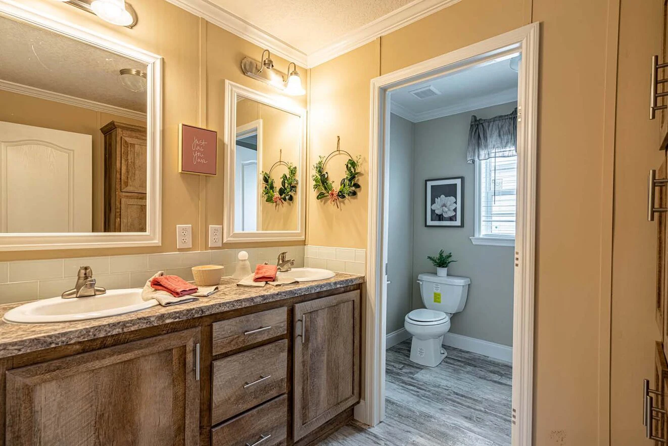 A bathroom with double sinks, wood cabinets, wall mirrors, and a separate toilet area with a window and decor.
