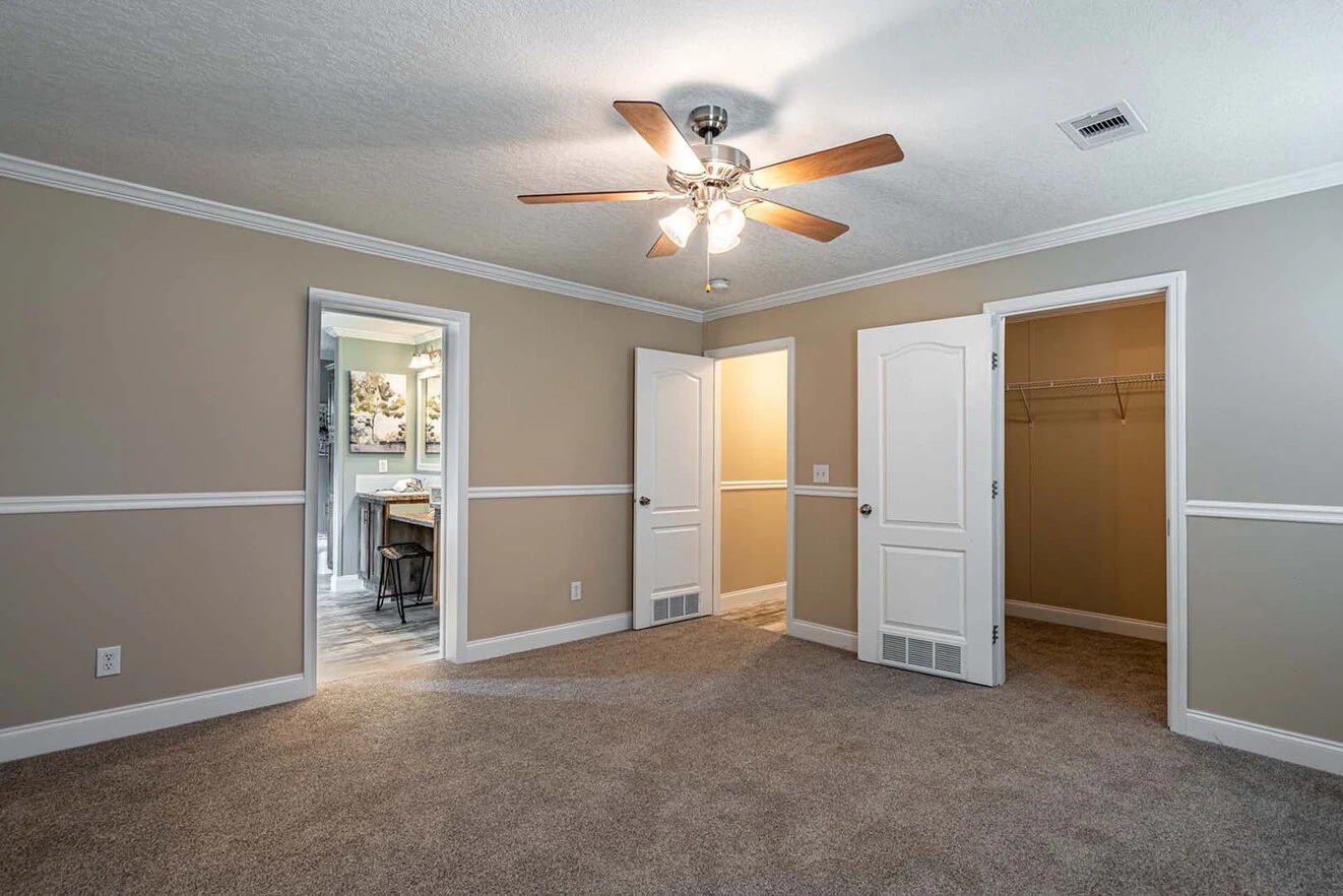 Empty carpeted bedroom with beige walls, ceiling fan, two open doors, one leading to a bathroom, and one to a walk-in closet.