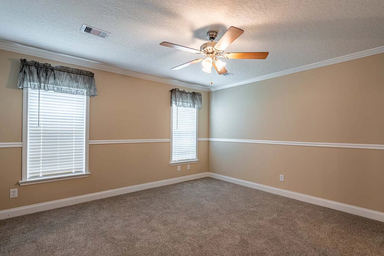 Empty beige-carpeted room with two windows, white blinds, short curtains, a ceiling fan with lights, and beige walls with white trim and chair rail molding.