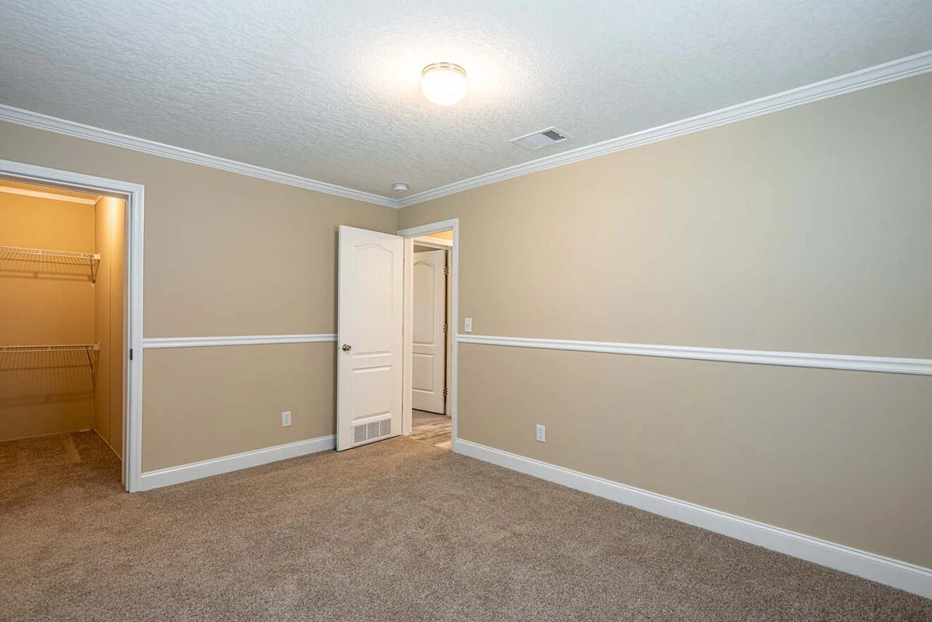 Empty beige bedroom with carpeted floor, white trim, a ceiling light, open door, and a walk-in closet with wire shelving.