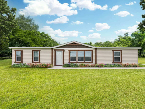A single-story manufactured home with beige siding, brown trim, and a white front door, surrounded by green lawn, trees, and a small flower bed in front.